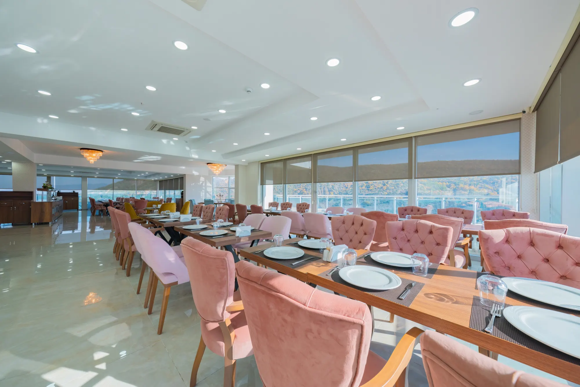 Geniş pencereli restoran salonu, düzenli kurulmuş masalar ve pembe kadife sandalyeler